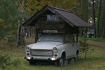 Synonym für die Anfahrt ist auf diesem Bild ein himmelblauer PKW Trabant mit einem hölzernen, dunklen Dachaufbau in Zeltform. An dessen Giebel in Fahrtrichtung befindet sich ein rechteckiges Fenster mit Gardine.