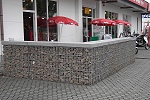 Eine etwa 15 Quadtratmeter große Sitzecke vor einem Imbiss wurde mit einer etwa 1 m hohen Gabionenwand eingefasst, die ebenfalls von einer etwa 10 cm dicken, grauen Betonplatte abgedeckt wurde. Umgeben ist die Wand von wabenförmigen Betonplastersteinen. Zu sehen sind außerdem kleine rote Sonnenschirme, zwei Angebotsaufsteller und der Eingangsbereich zum Imbiss.