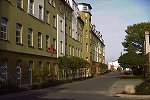 Zu sehen ist eine Straße mit einer olivgrünen historischen Häuserzeile, ein Gehweg davor und ein Grünstreifen, bevor die Straße beginnt. Im Grünstreifen wurden zwischen zweiflammigen roten Straßenleuchten jeweils vier Laubbäume gepflanzt. Diese sind etwa 4 m hoch.