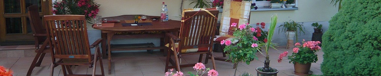 Eine dunkle, hölzerne Sitzgruppe lädt auf einer überdachten Terrasse inmitten von lachsfarbenen Blumen zum Verweilen ein. Die Sitzgruppe besteht aus einer Bank, zwei hohen Klappstühlen und einem Tisch.