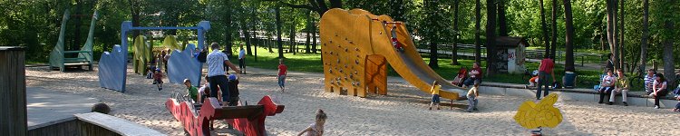 Ein großer Spielplatz mit überdimensionalen Tieren mitten in einem schattigen Waldstück läd zum Spielen ein. Auf Bänken rund um die Anlage verweilen zahlreiche Erwachsene, während die Kinder zum Beispiel auf orangen Elefanten und blauen Saurieren rutschen und schaukeln.